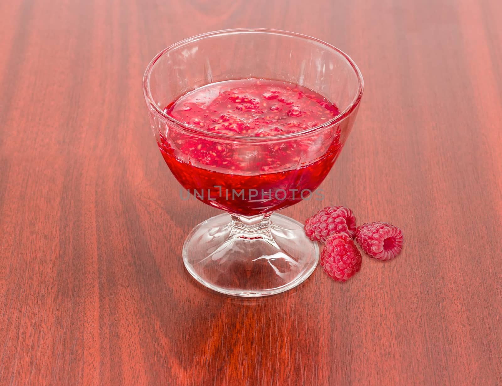 Raspberry jam in the glass dessert stem bowl and several fresh berries beside on a red wooden table
