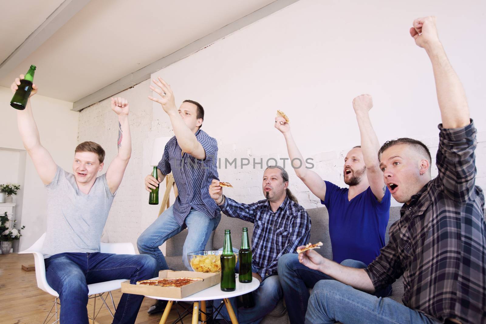 Male friends sports fans watching winning football match on TV at home celebrating winning goal huddled on couch shouting excited sharing snacks drinking beer