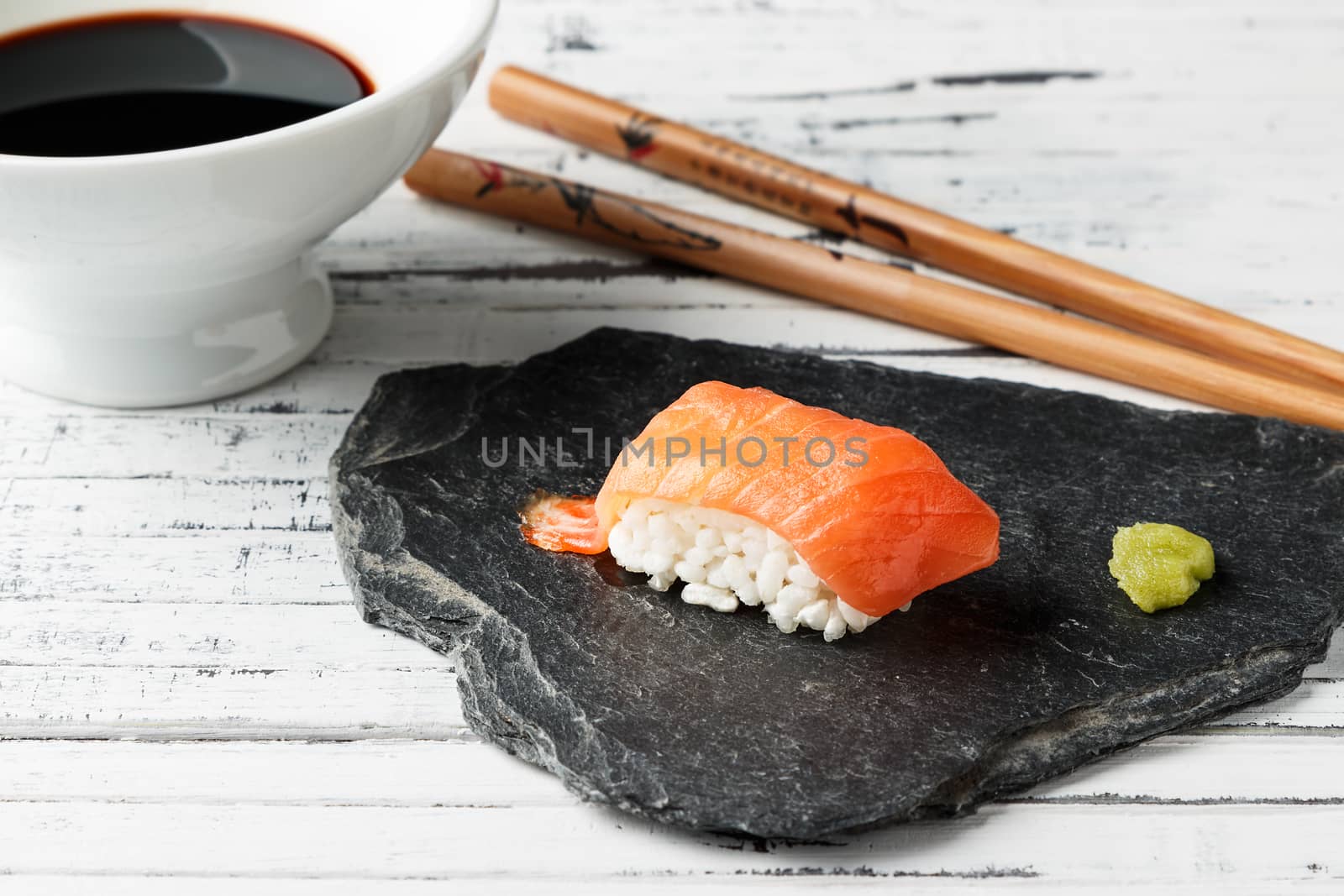 Smoked salmon Nigiri with wasabi paste on slate stone. Chopsticks and bowl with soy sauce in the background on old white wood. Raw fish in traditional Japanese sushi style. Horizontal image.