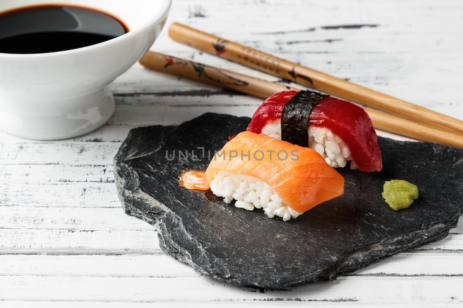 Smoked salmon Nigiri with wasabi paste and red tuna nigiri on slate stone. Chopsticks and bowl with soy sauce in the background on old white wood. Raw fish in traditional Japanese sushi style. Horizontal image.