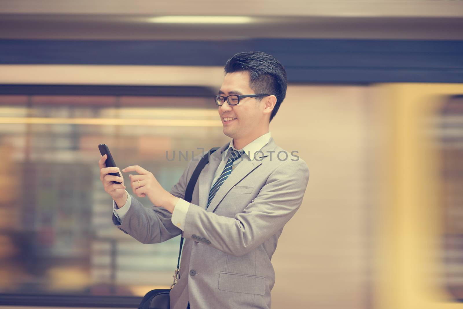 Asian businessman using smart phone in subway station, train passing by at the background.