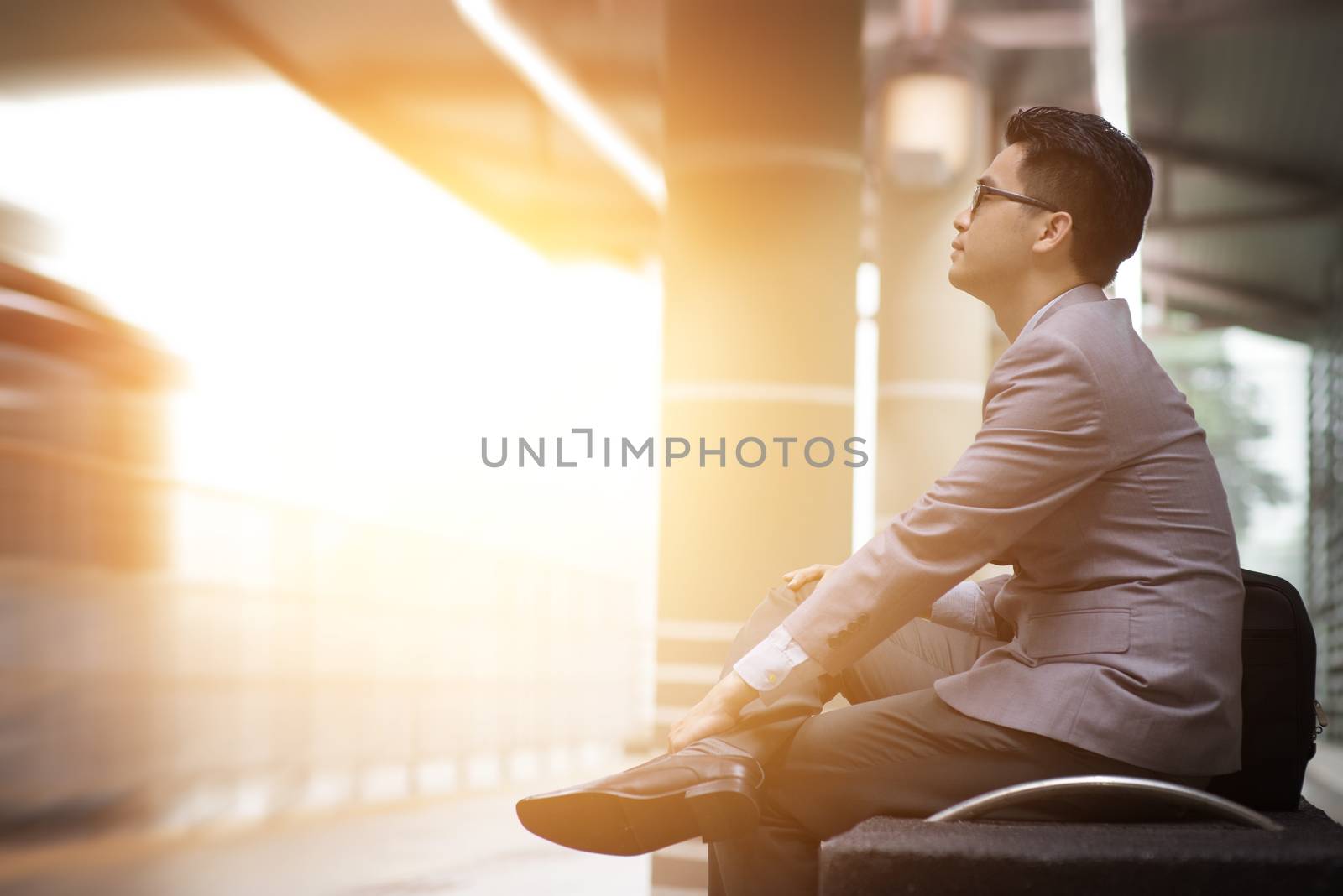 Asian businessman sitting at railway station, waiting for train, beautiful sun light at background.