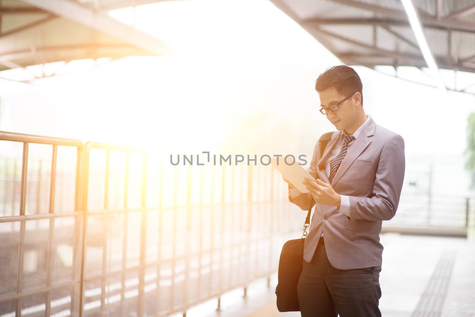 Asian businessman using digital tablet pc at railway platform, with beautiful sun flare at background.