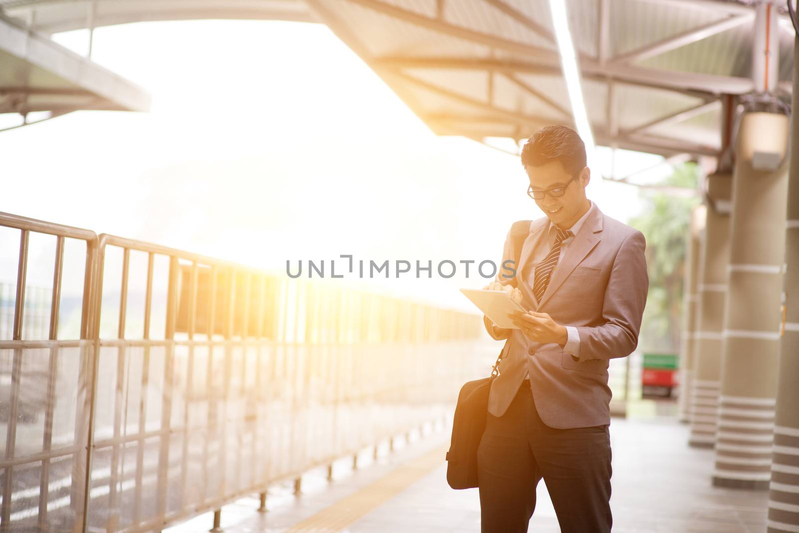 Asian businessman using digital tablet pc at metro transit platform, with beautiful sun flare at background.