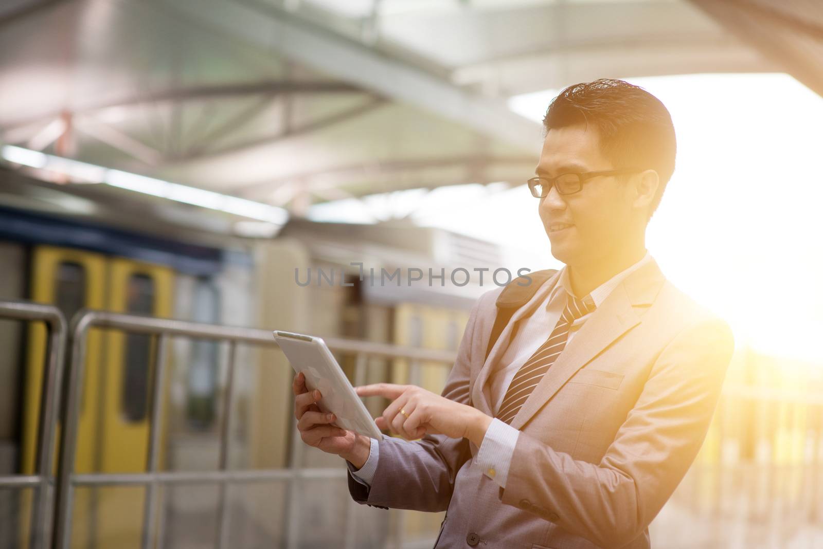 Asian businessman using digital tablet pc while waiting commuter, with beautiful sun flare at background.