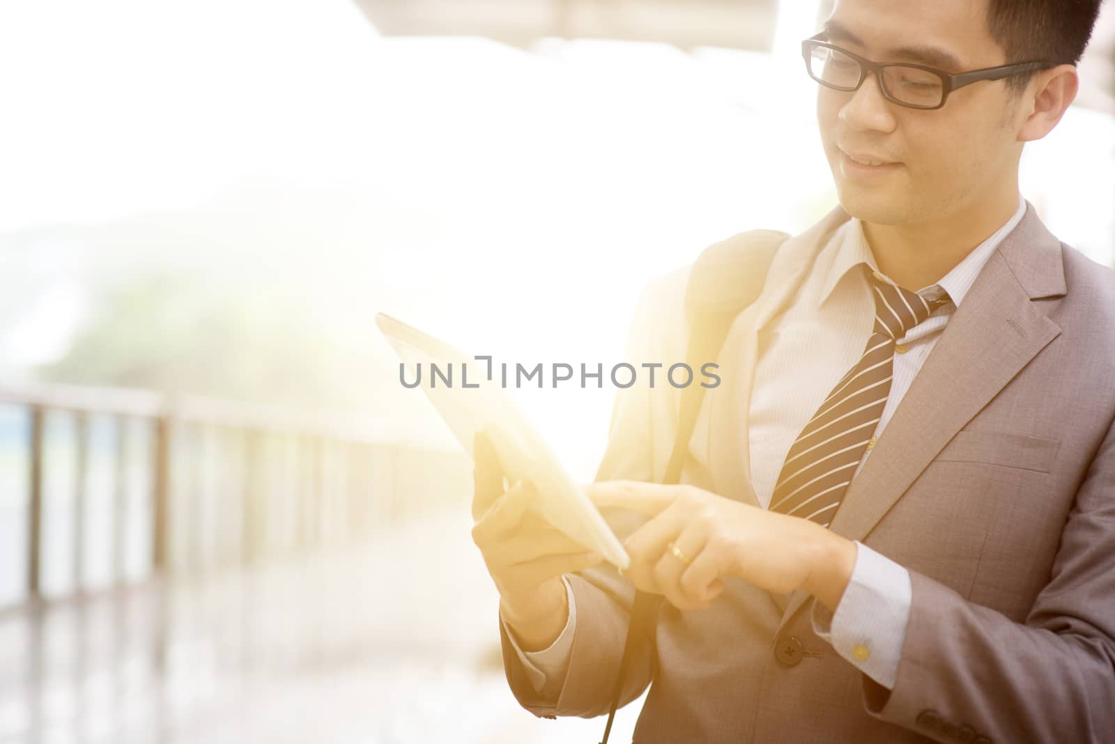 Asian businessman using digital tablet pc while waiting train, with beautiful sun flare at background.