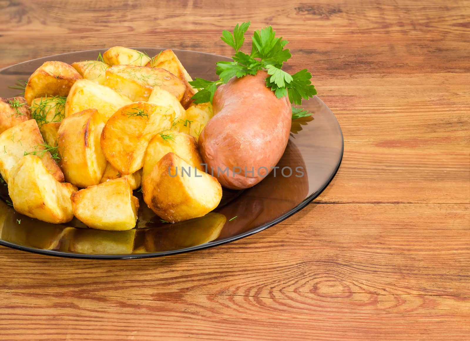 Fragment of the dark glass dish with fried potatoes sprinkled by chopped dill, fried wieners and twig of parsley closeup on a surface of old wooden planks
