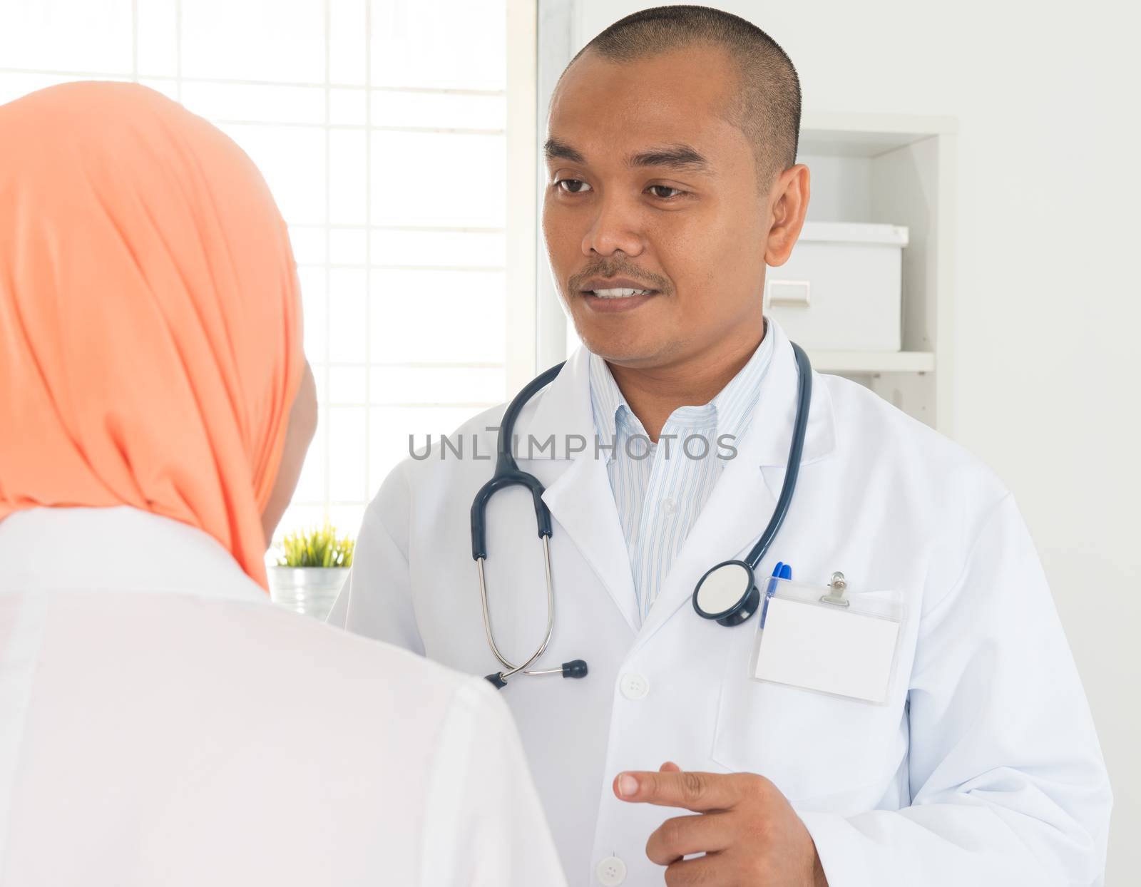 Male medical doctor talking with nurse inside hospital. Southeast Asian Muslim people.