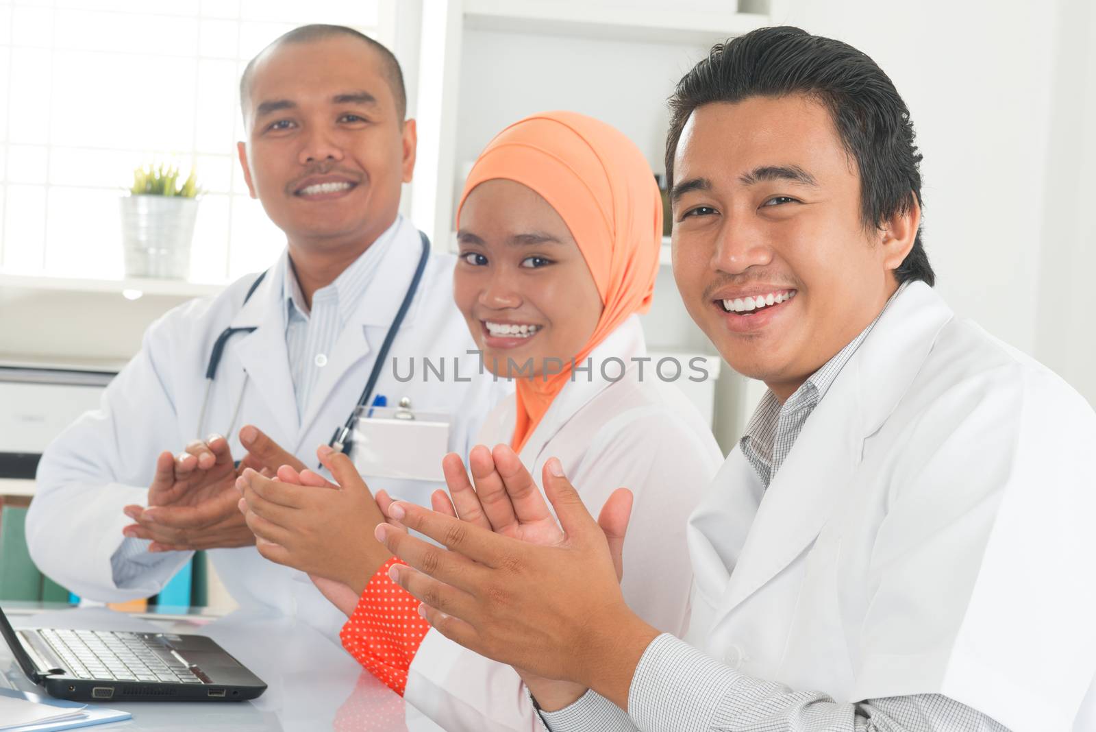 Medical doctors clapping hands during meeting on desk inside hospital room. Southeast Asian Muslim medical people.