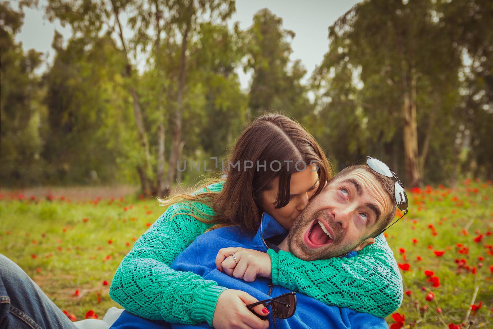 Young couple sitting on the grass in a field of red poppies. The girl kiss the guy while he has a silly smile.