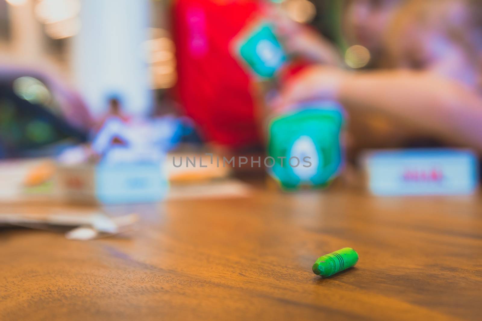 Green color pastel crayon on brown wooden table. Children playing in blurry background. Close up selective focus.