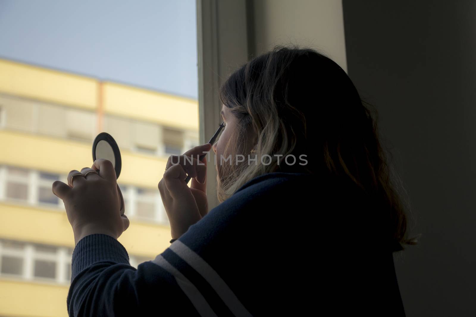 Back view of silhouette of a woman putting on makeup in front of a window.