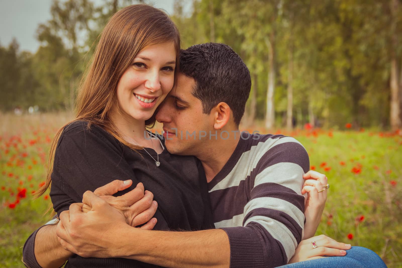 Young couple sitting on the grass in a field of red poppies and smiling at the camera.