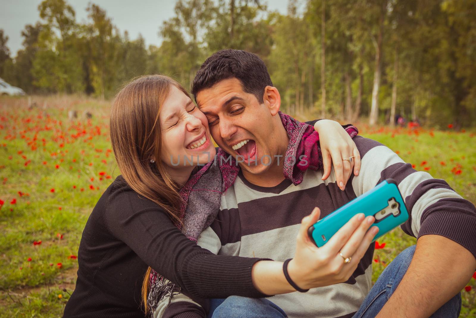 Couple doing silly and funny faces while taking selfie picture with their mobile phone in field of red poppies.