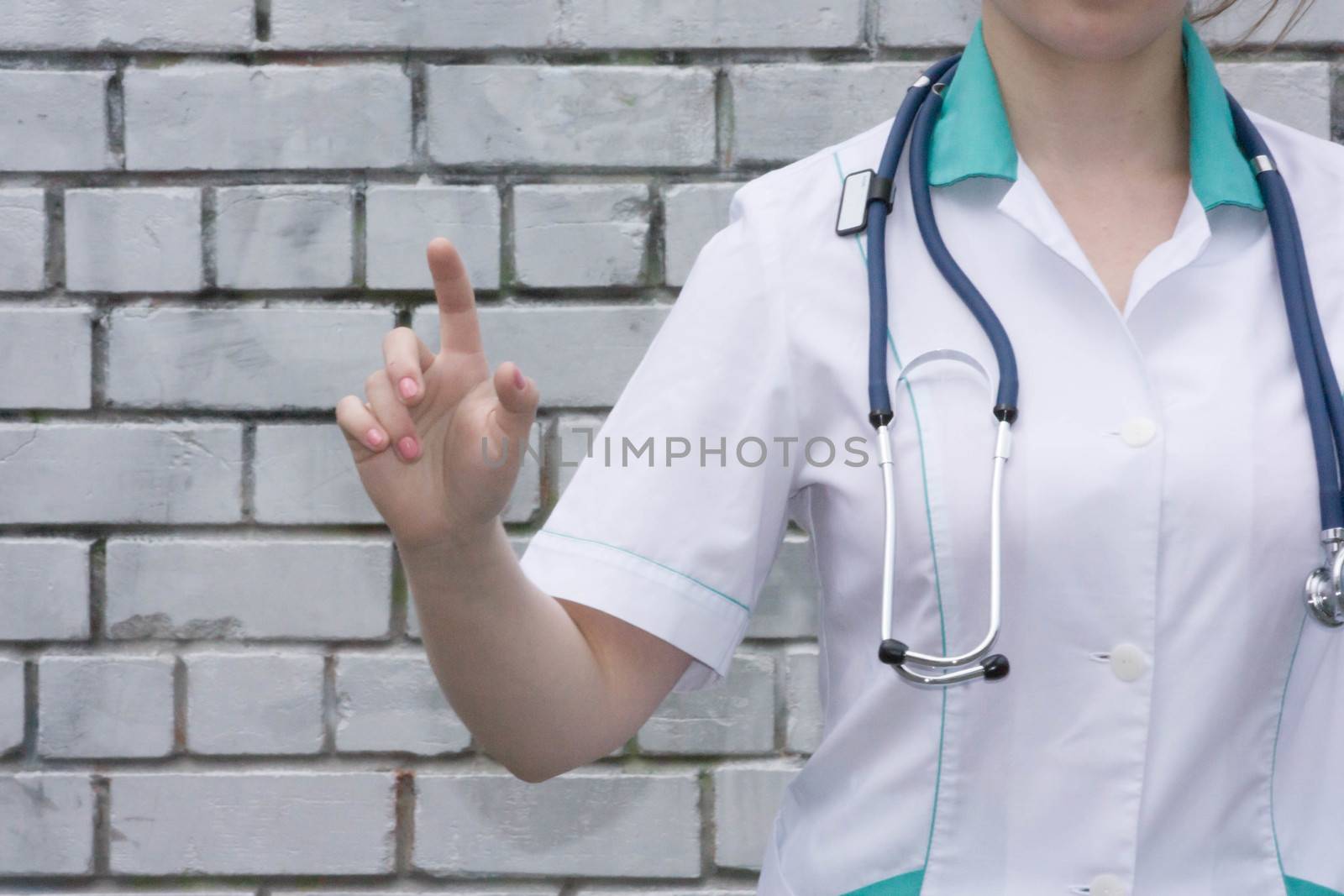 The doctor's concept. Girl in medical uniform with a stethoscope near a brick wall. Holds a finger finger forward. Presses. Photo for your design