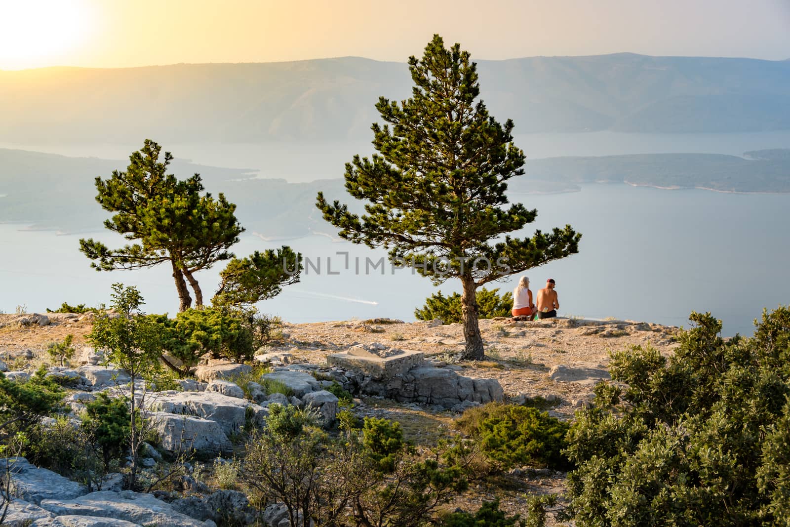 A mature couple sitting on a large rock overlooking the seashore at sunset and looks at the horizon by asafaric