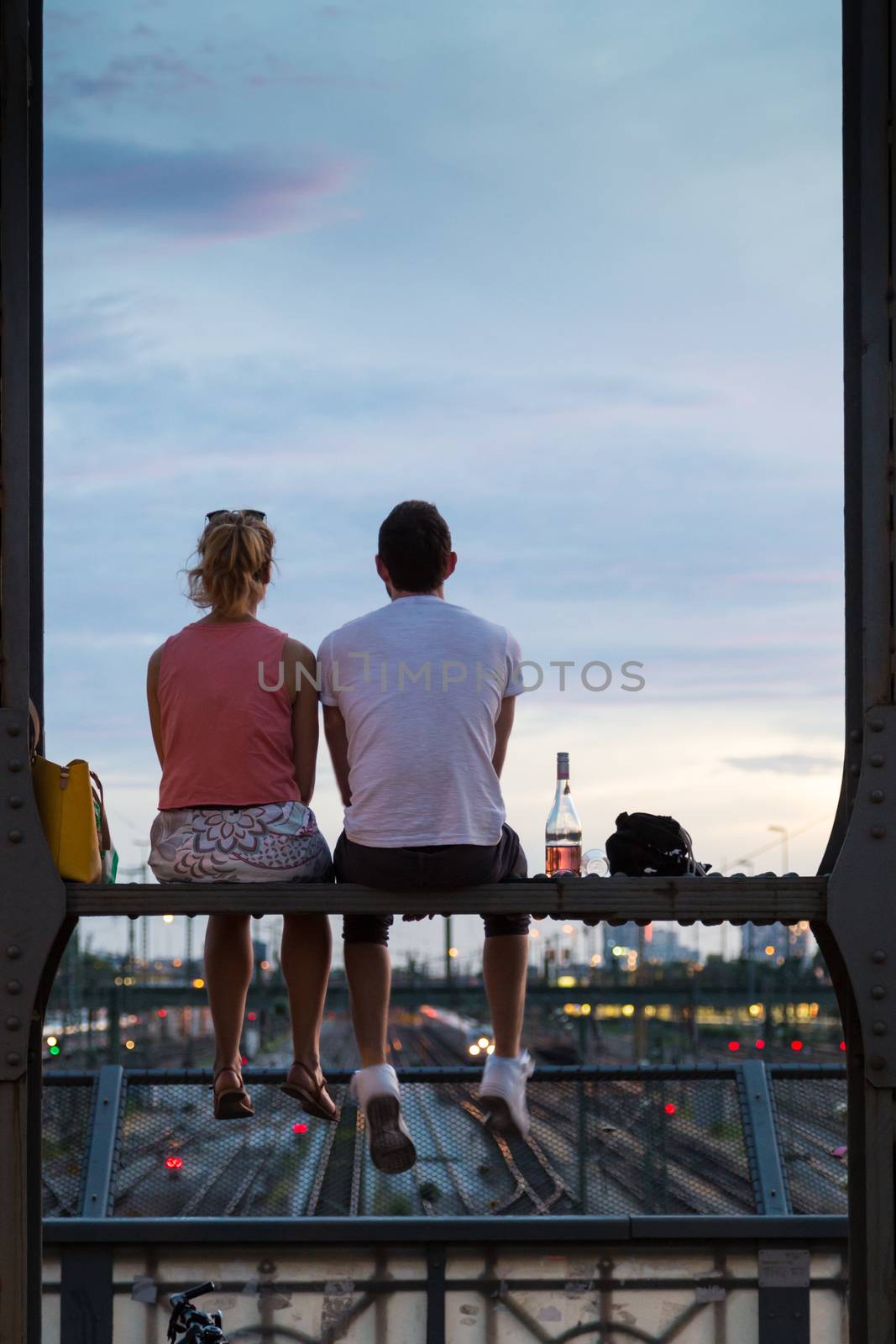 Young couple in love on romantic date sitting on railway bridge with a bottle of wine and watching the sunset over Munich urban city center. Just the two of us.