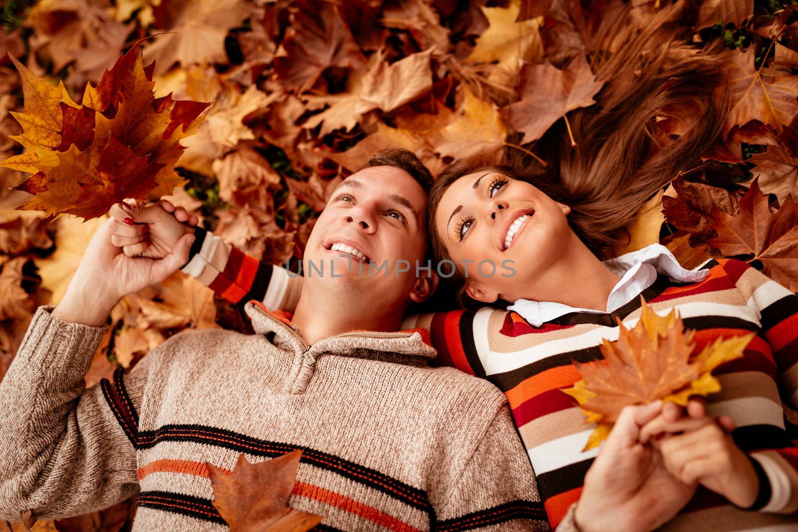 Beautiful young couple in sunny forest in autumn colors. They are lying on the ground covered with leaves and enjoying. 