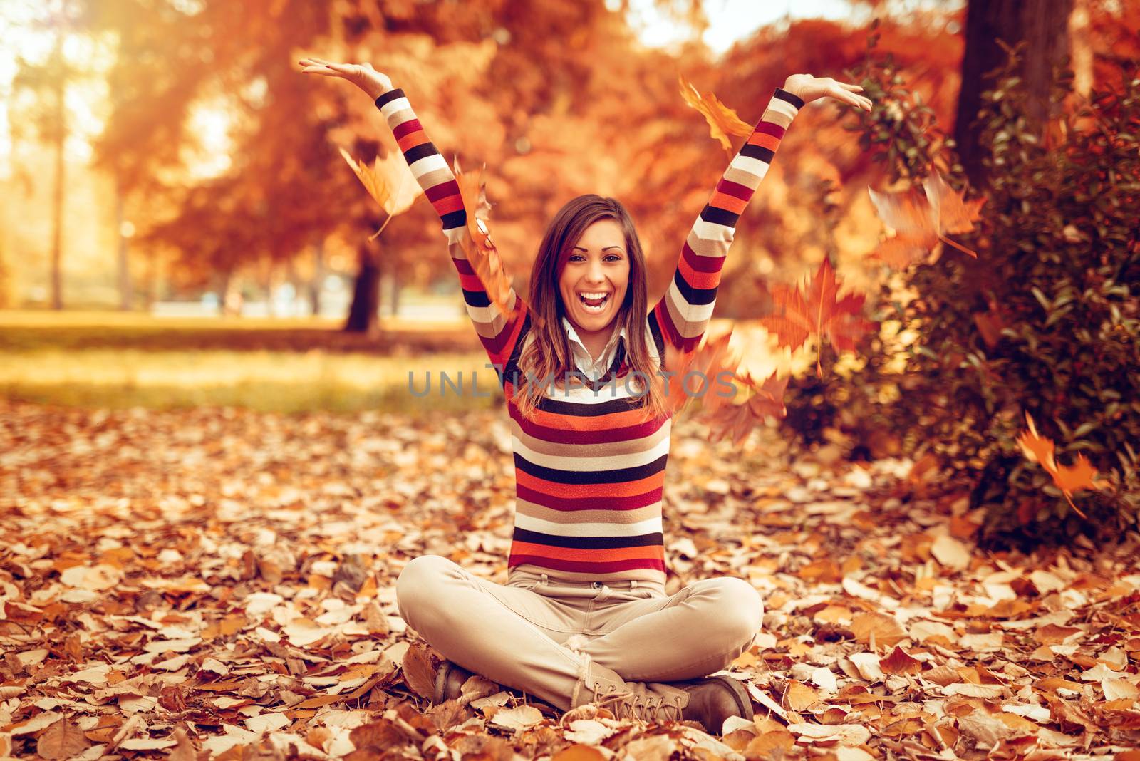 Cute young woman enjoying in sunny forest in autumn colors. She is sitting on the ground covered with leaves and having fun.