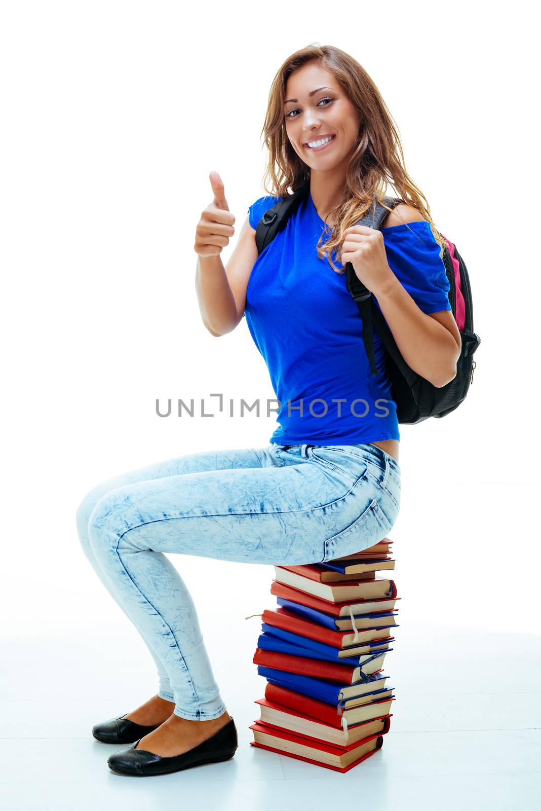 Beautiful smiling teenage girl with school bag sitting on stack of books and looking at camera with thumb up.