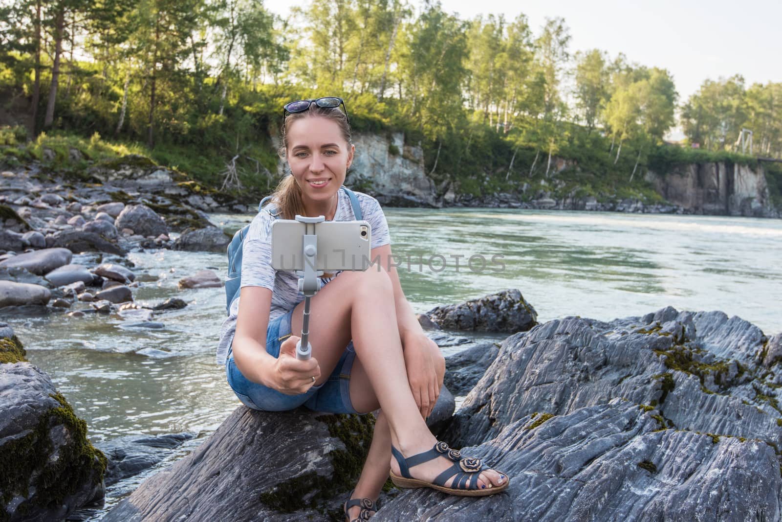 Woman taking selfie on mobile phone with stick on mountain river background