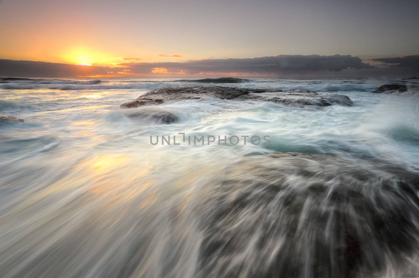 Ocean flowing rapidly over exposed rocks as the sun's glow peeks from behind clouds on the horizon.  Location, Bungan Beach, nortnern beaches Sydney Australia