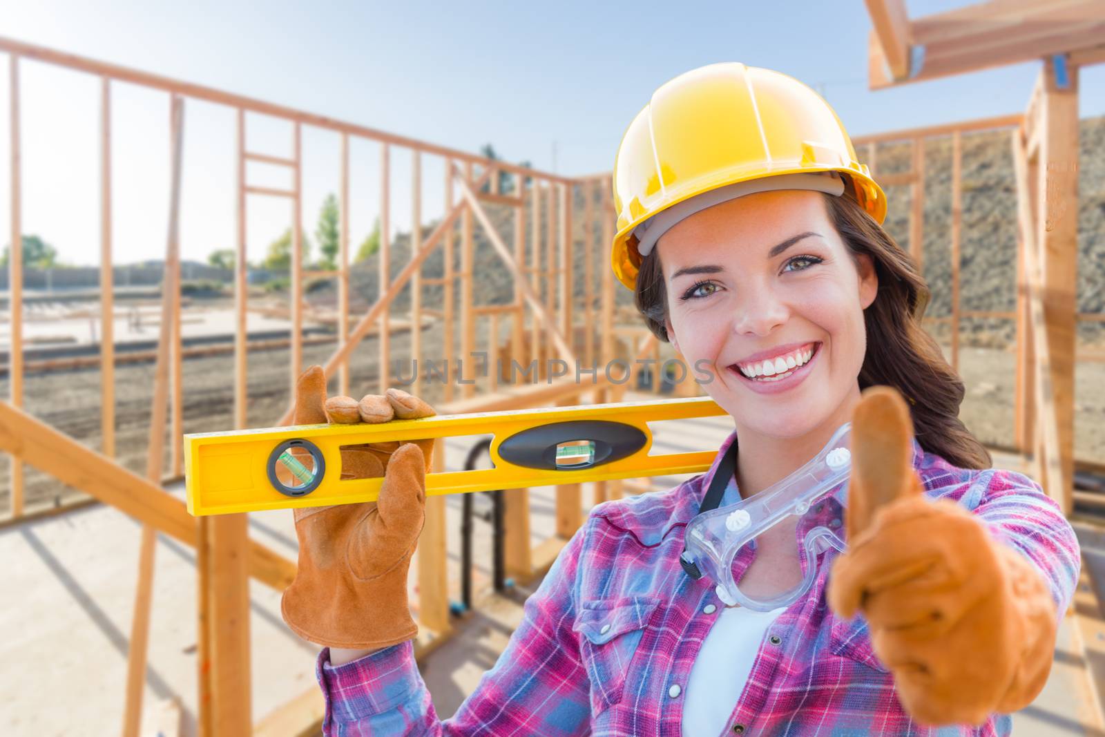 Female Construction Worker with Thumbs Up Holding Level Wearing Gloves, Hard Hat and Protective Goggles at Construction Site.