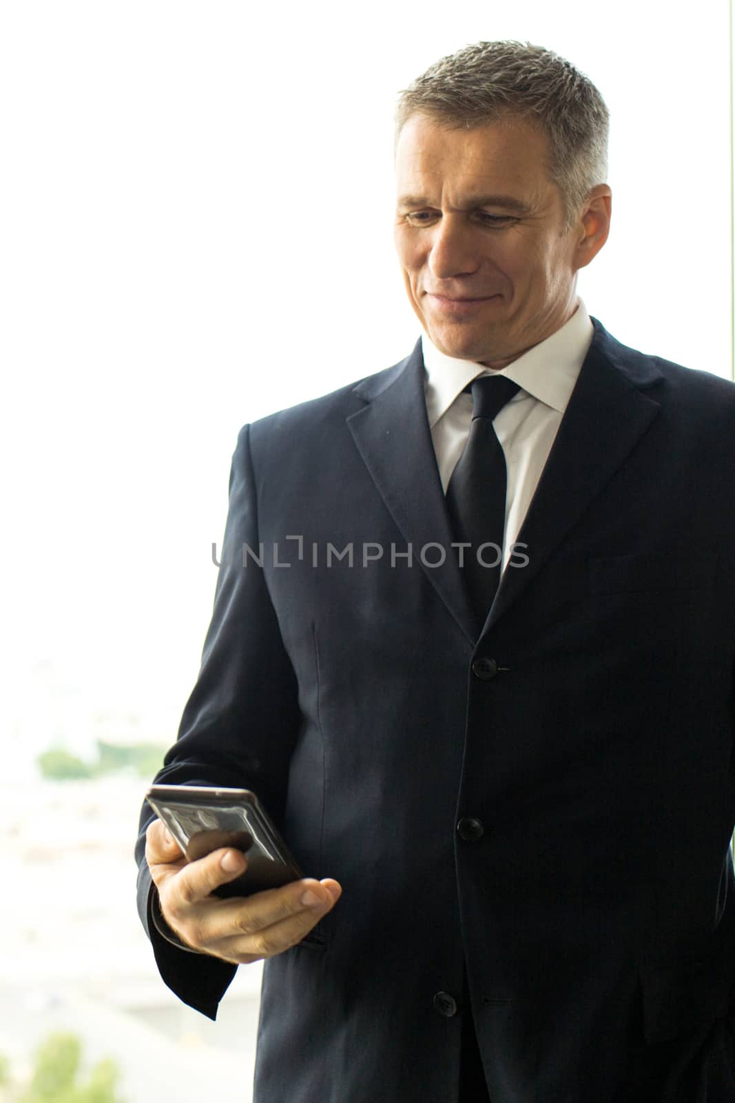 Mature businessman using smartphone standing near office window with view of city