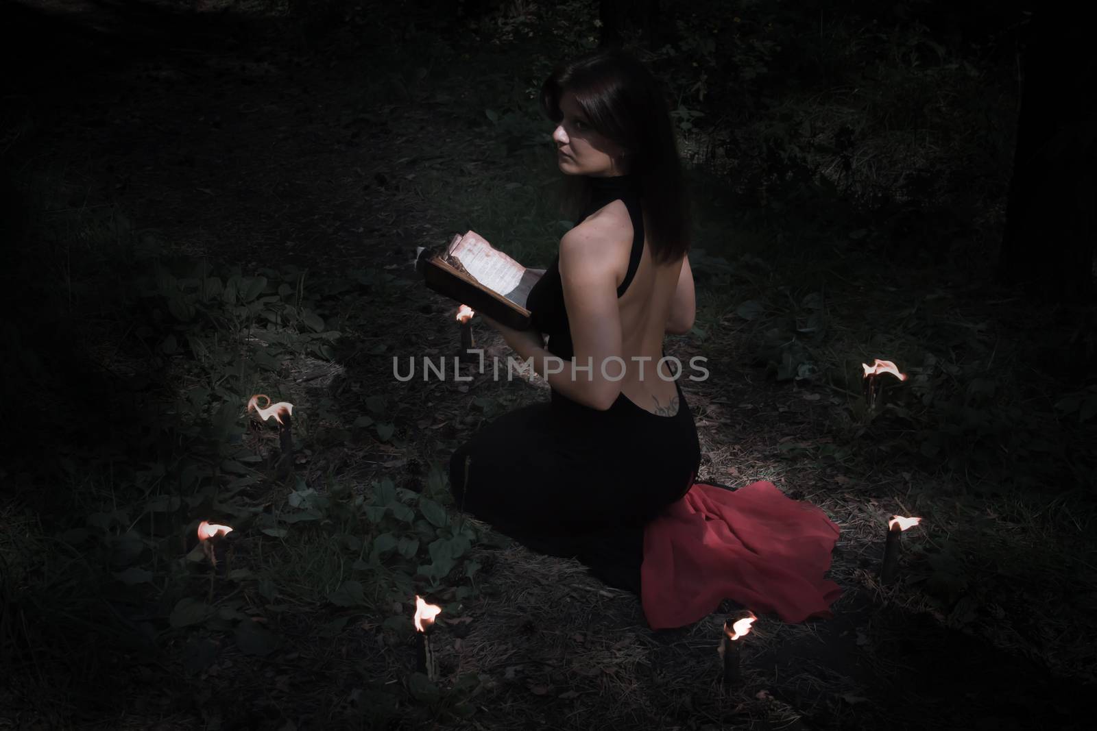 Halloween concept. Fairy in black. In the witchy circle. With a book of spells in hand. Looks up. Among woods and trees. With a beautiful back