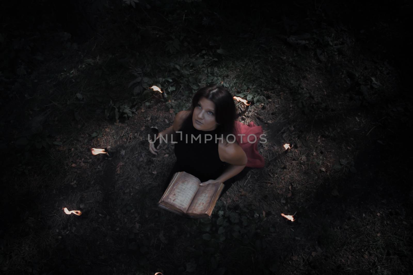 Halloween concept. Fairy in black. In the witchy circle. With a book of spells in hand. Looks up. Among woods and trees. Lighted candles of fire. Witchcraft and magic