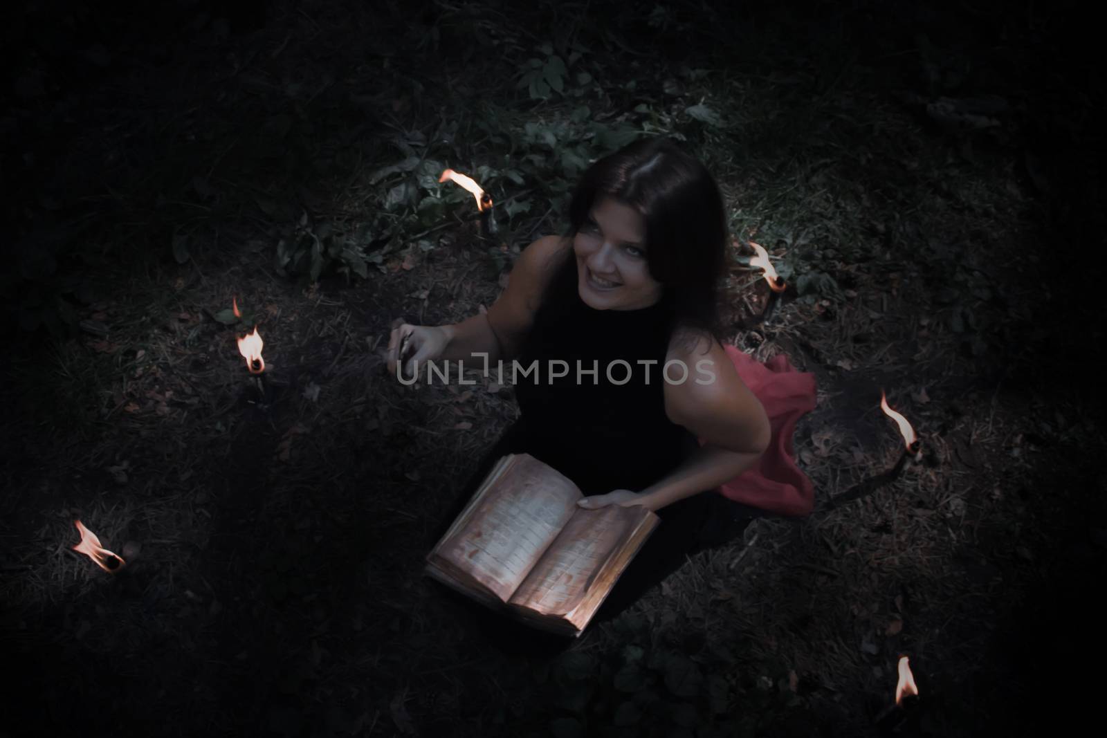 Halloween concept. Witch in black. In the witchy circle. With a satanic book in his hands. Looks up. Among woods and trees. Lighted candles of fire. Witchcraft and magic. Photo for your design.