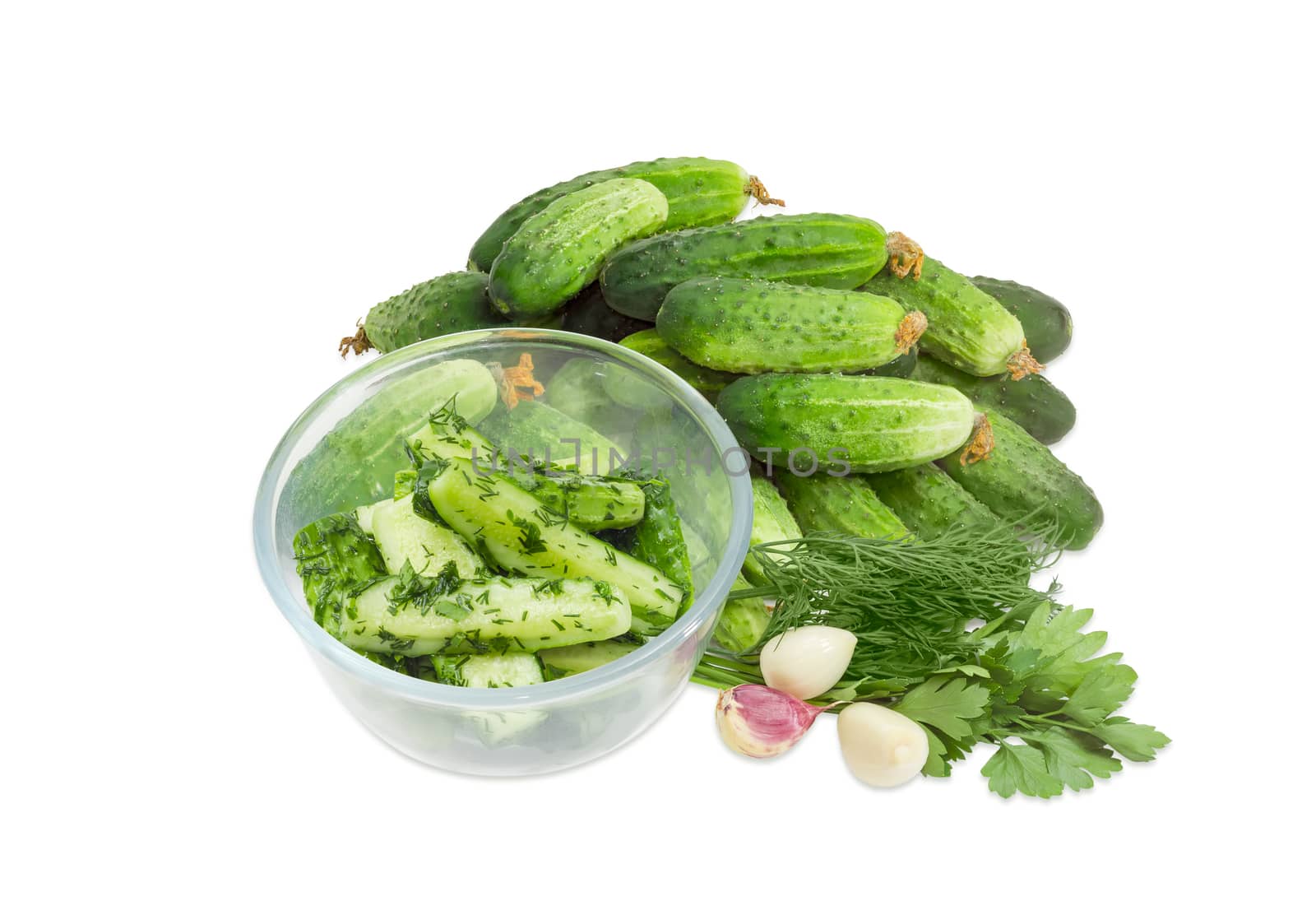 Lightly salted cucumbers in the glass bowl against of a pile of the freshly picked out cucumbers, parsley, dill and garlic on a white background
