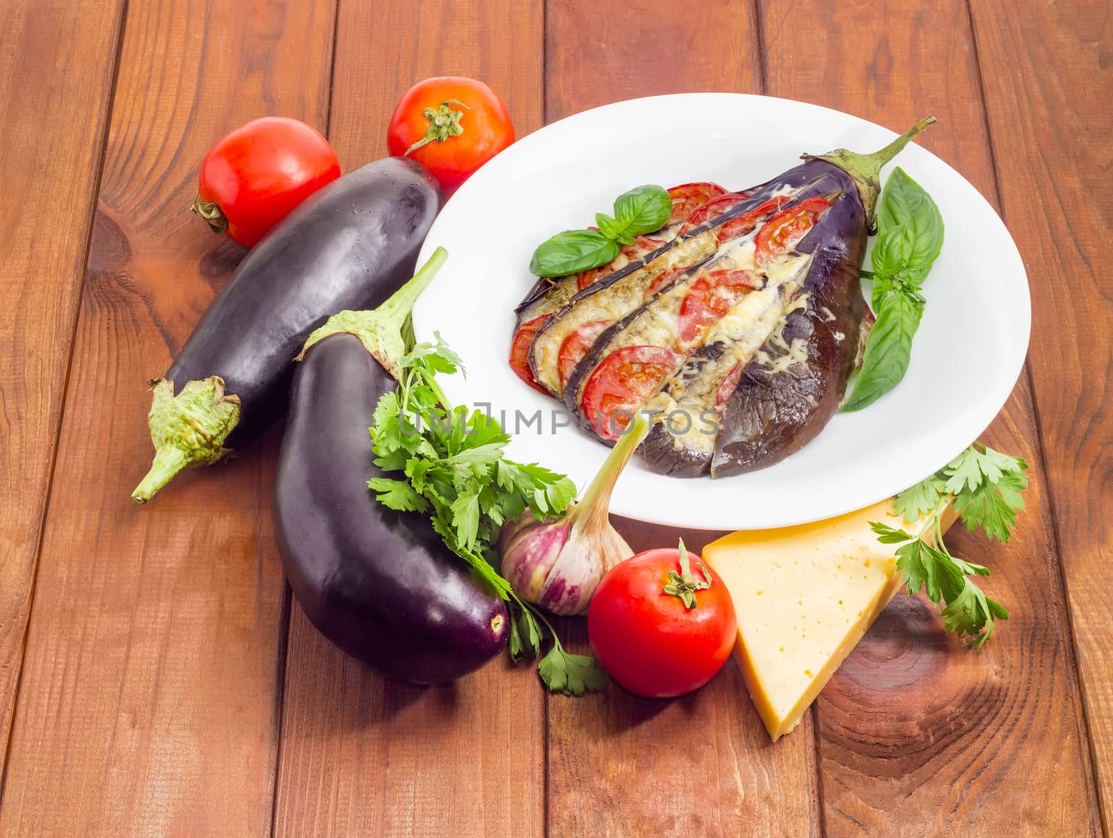 Baked eggplant stuffed with vegetables and cheese decorated with basil leaves on white dish and ingredients for its cooking beside on a surface of a dark wooden planks
