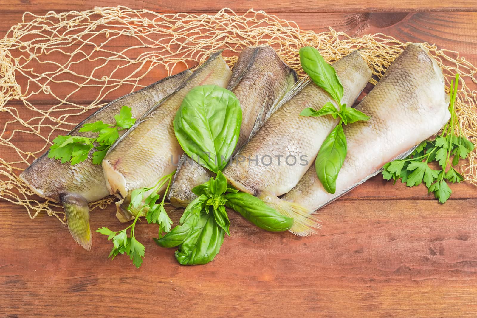 Several uncooked carcasses of the notothenia fish without of head and tail and twigs of basil and parsley on the fishing net on a dark wooden surface
