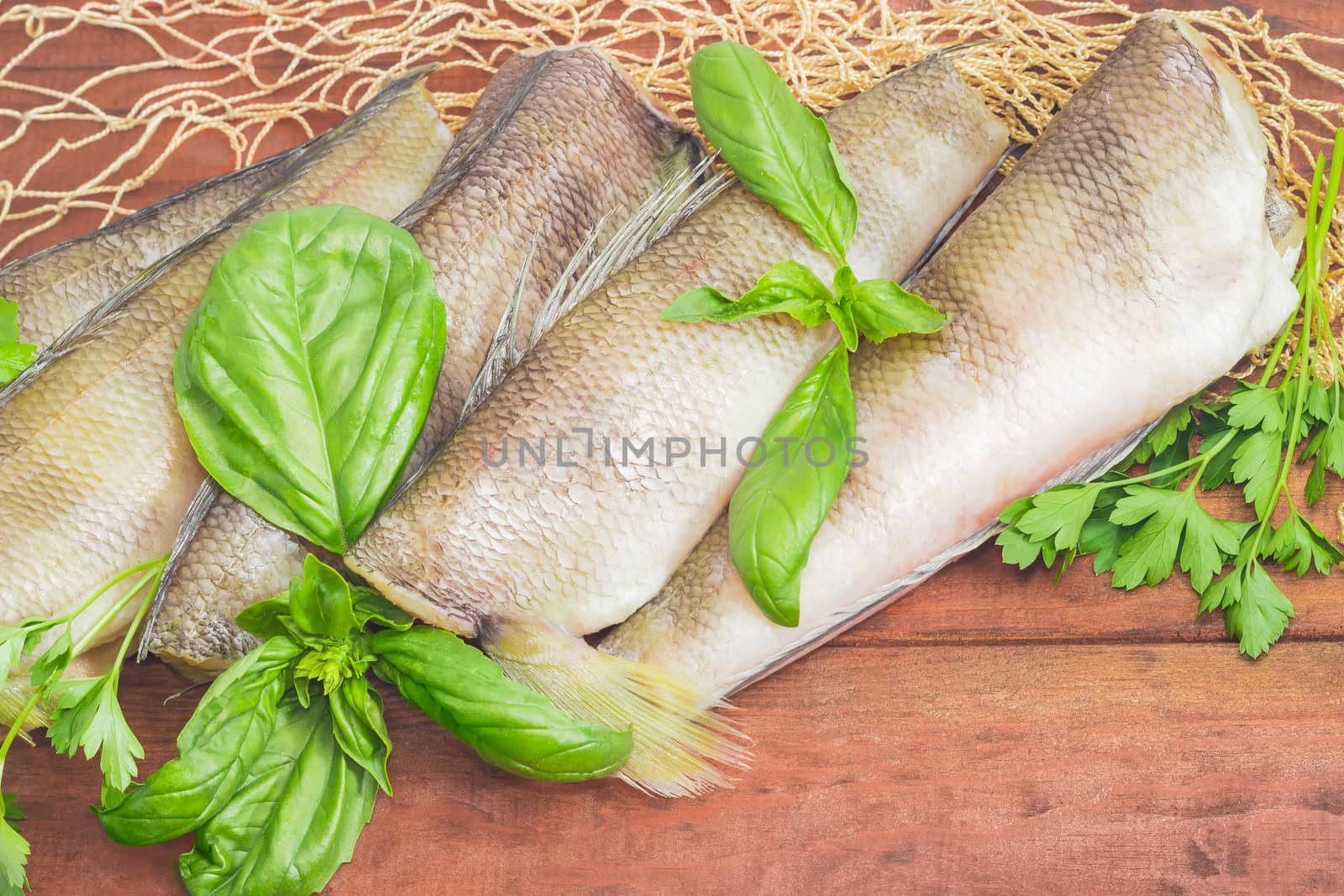 Several uncooked carcasses of the notothenia fish without of a heads and tails and twigs of basil and parsley closeup on the fishing net on a dark wooden surface
