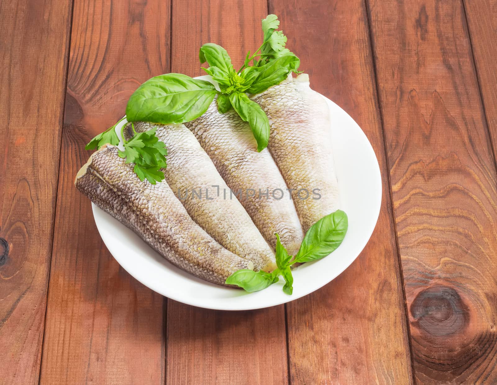 Uncooked carcasses of the notothenia fish without of a heads and tails and with peeled scales and prepared for cooking, twigs of basil and parsley on the white dish on a dark wooden surface
