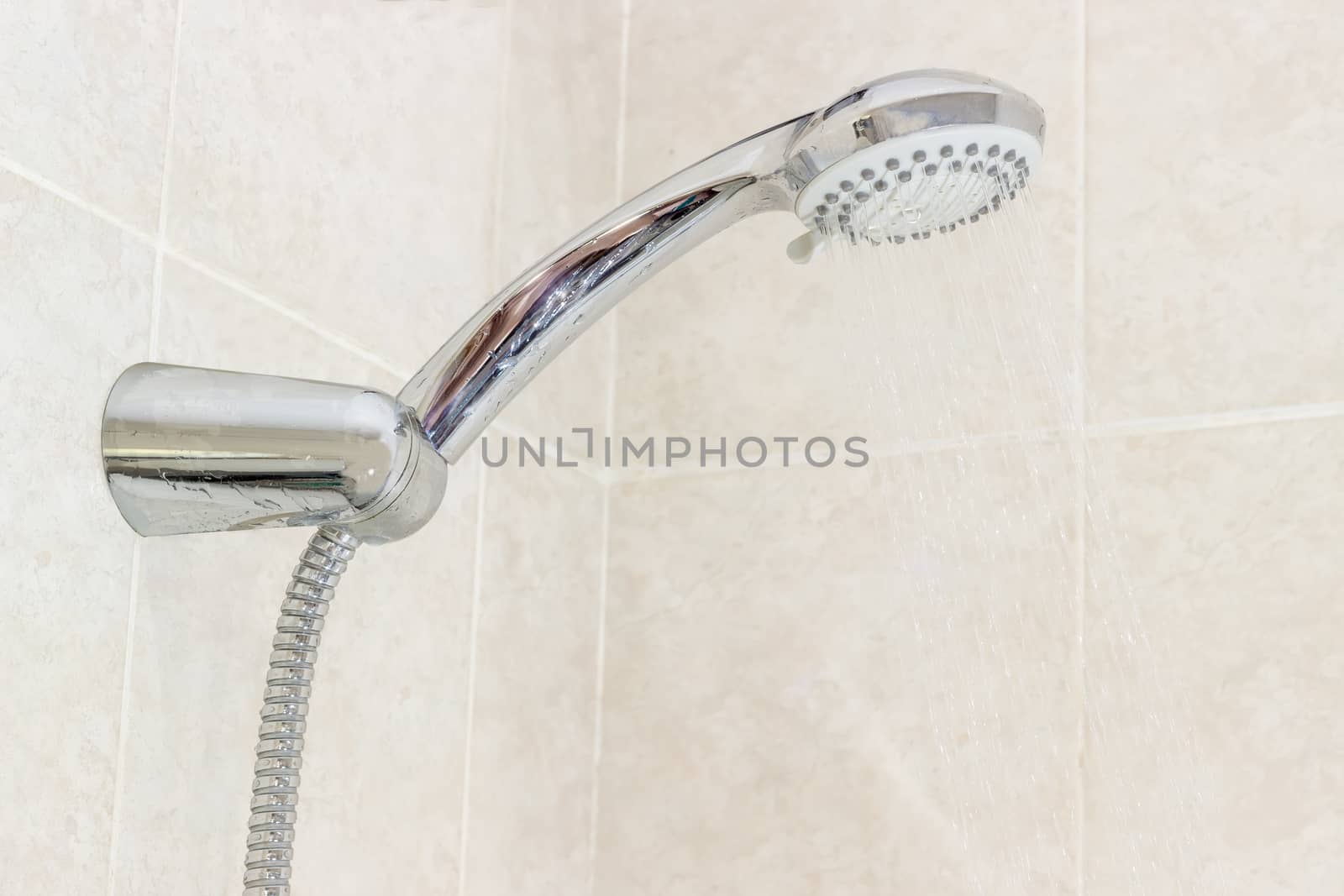 Shower head with metal shower hose in holder and water jets closeup on a background of a wall with beige tiles
