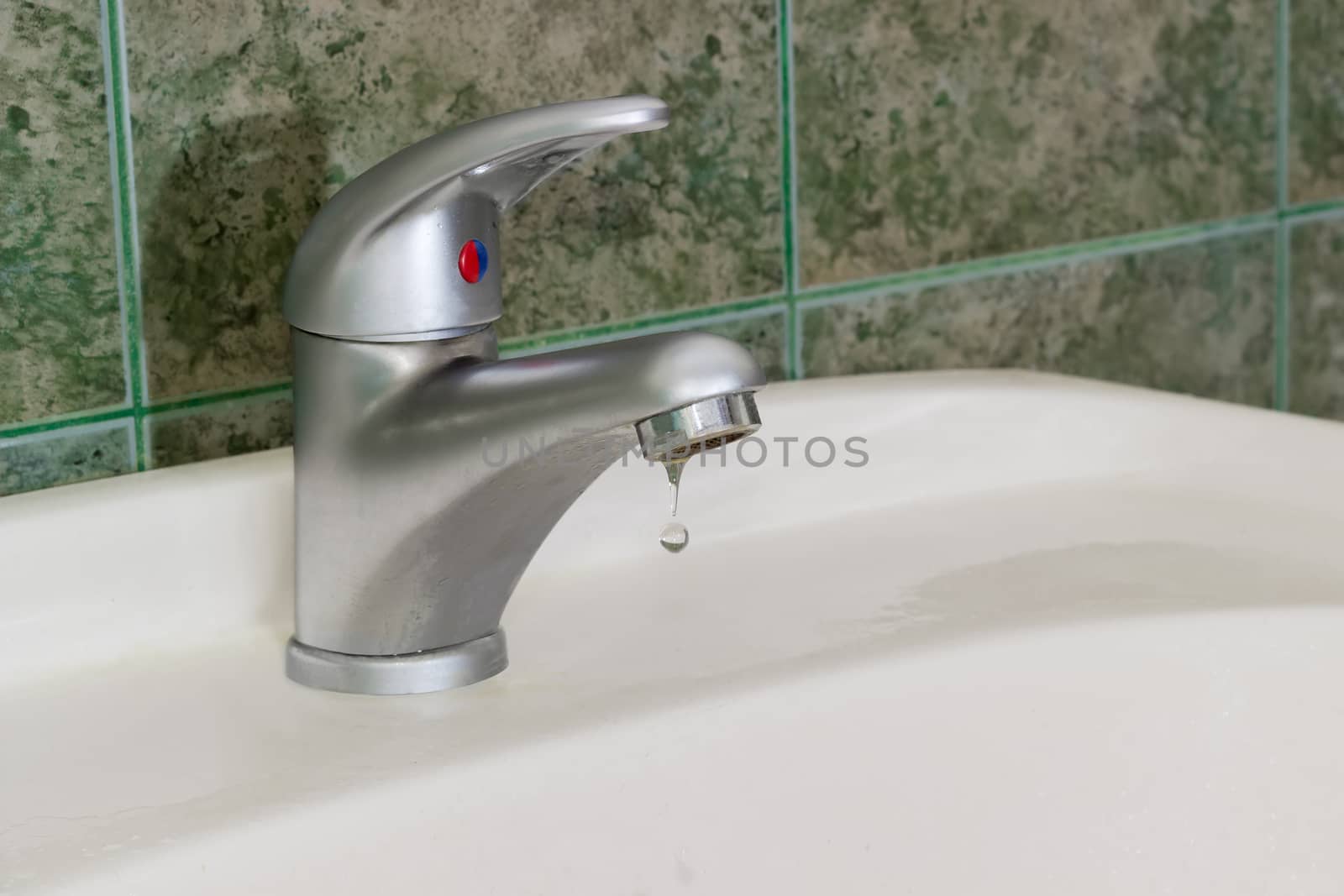 Drop of water from the single handle mixer tap mounted on a wash basin on background of a wall with green tiles
