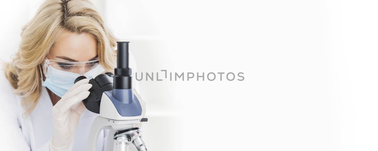 Female doctor in protective glasses and mask looking through a microscope, studio shot isolated on white background