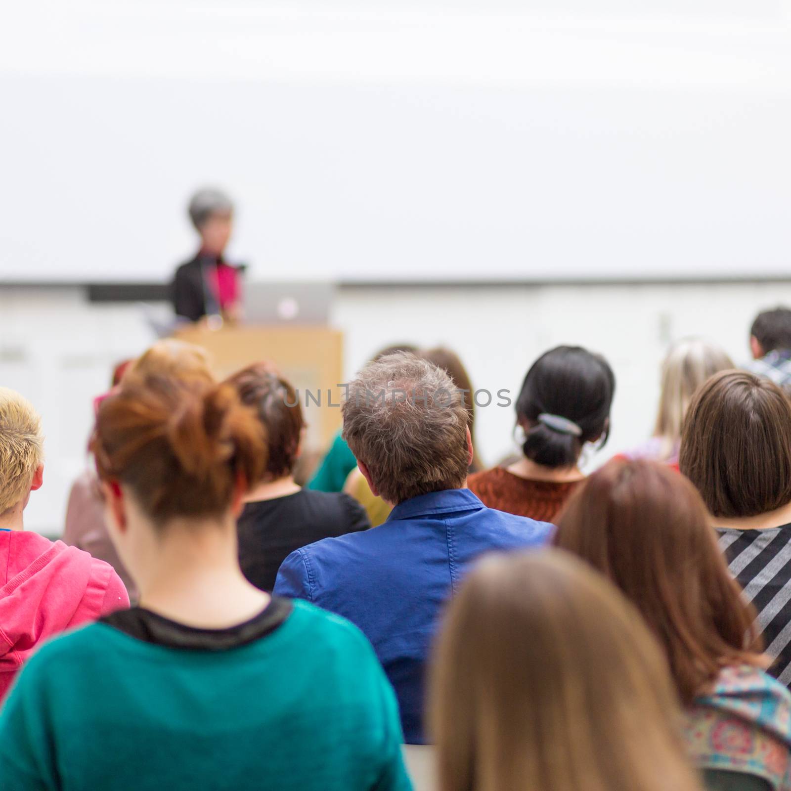 Business and entrepreneurship symposium. Female speaker giving a talk at business meeting. Audience in conference hall. Rear view of unrecognized participant in audience.