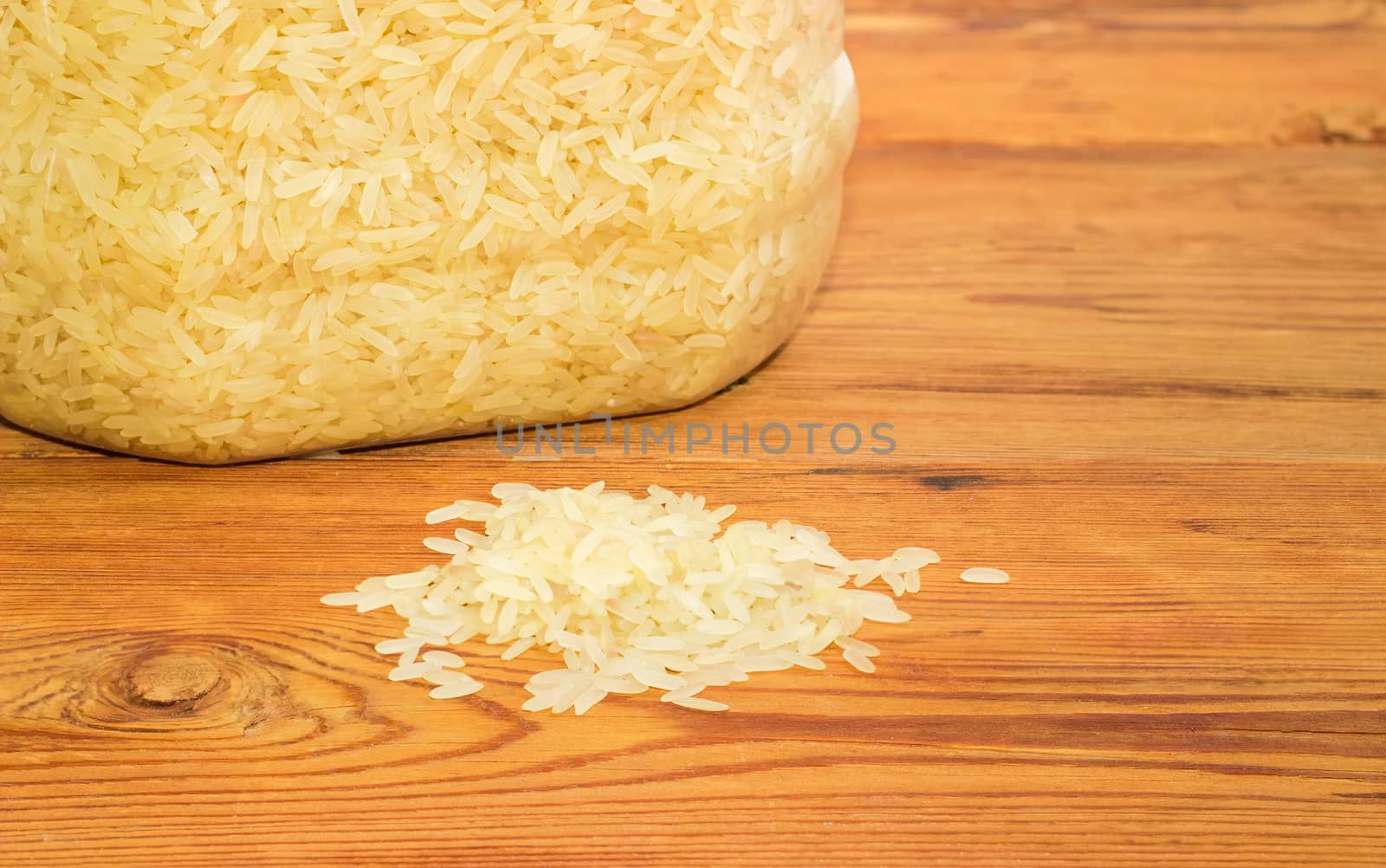 Small pile of the uncooked rice closeup against of part of the transparent plastic container with rice on the surface of the old wooden planks
