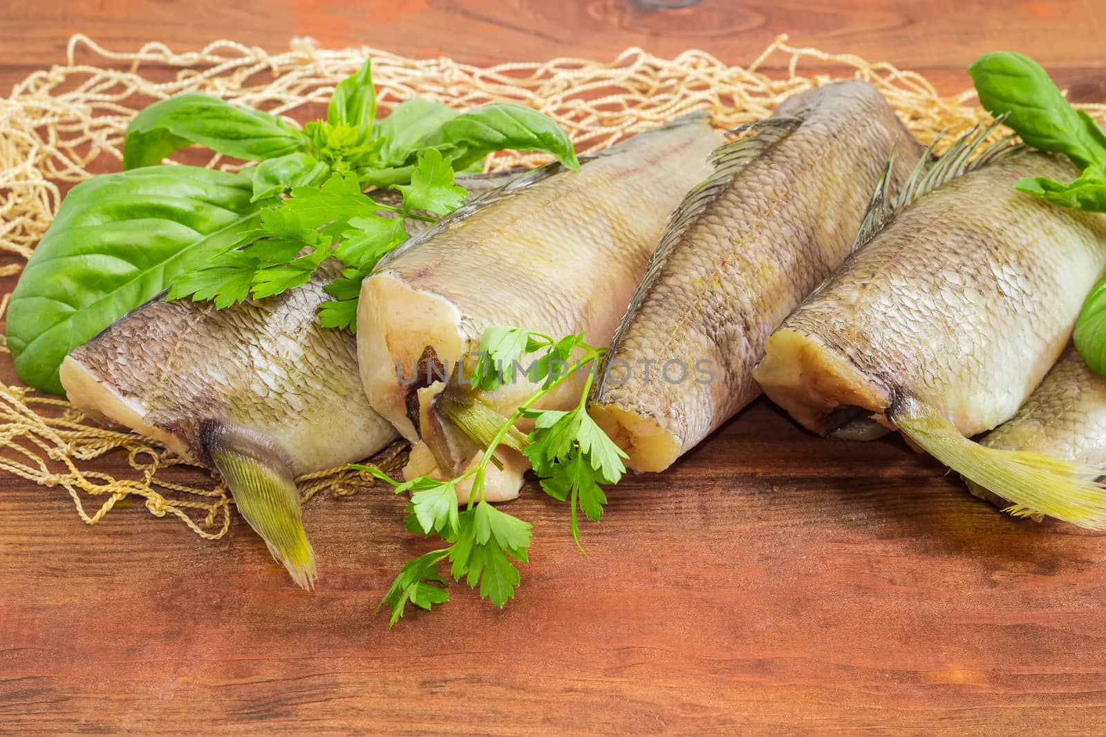 Several uncooked carcasses of the notothenia fish without of a heads and tails and twigs of basil and parsley closeup on the fishing net on a dark wooden surface
