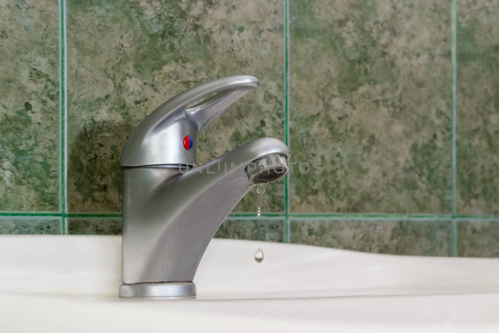 Dripping water from the single handle mixer tap mounted on a wash basin on background of a wall with green tiles
