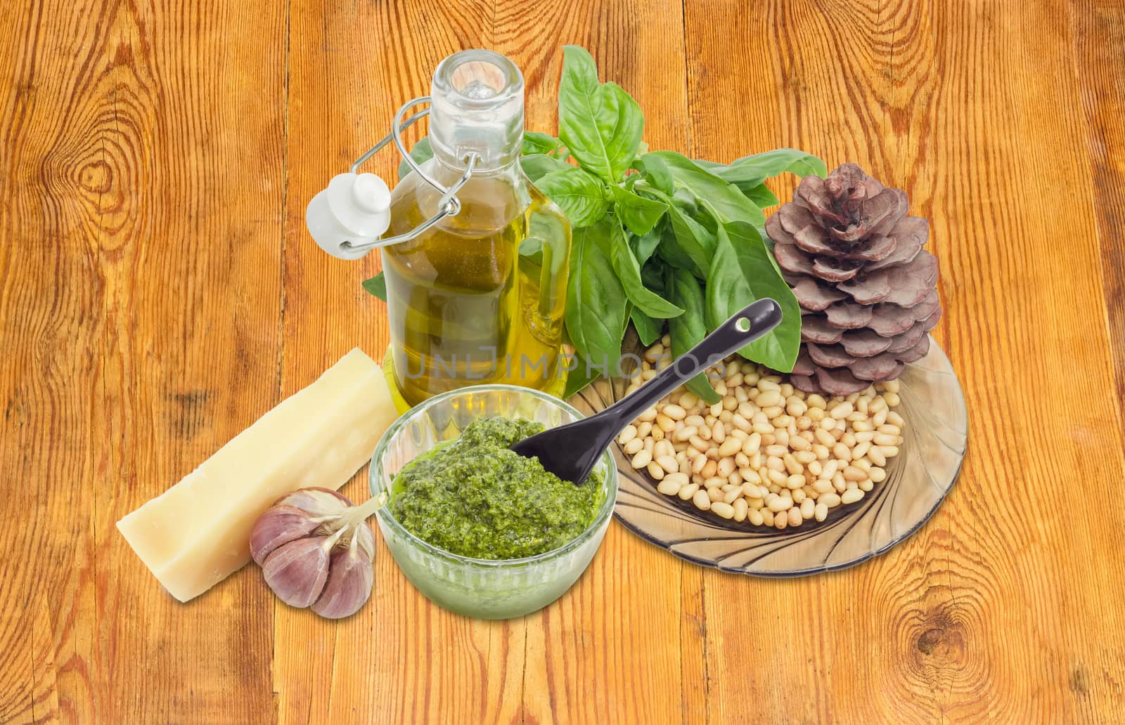 Sauce basil pesto in the small glass bowl with small black ceramic spoon on a background of ingredients for its preparation and pine cone on a surface of an old wooden planks
