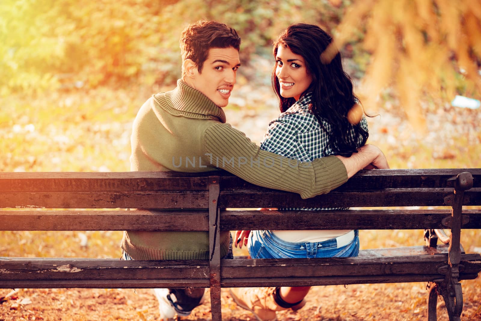 Beautiful lovely couple sitting on the bench and enjoying in sunny park in autumn colors. Looking at camera. Rear view.