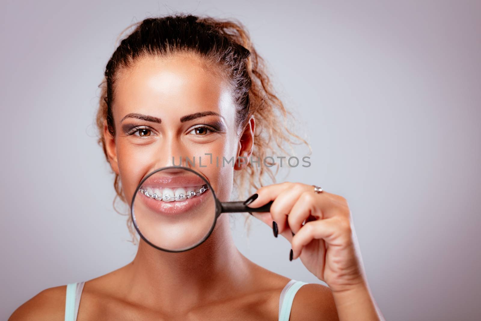 Smiling girl with braces on teeth behind magnifying glass. Looking at camera.