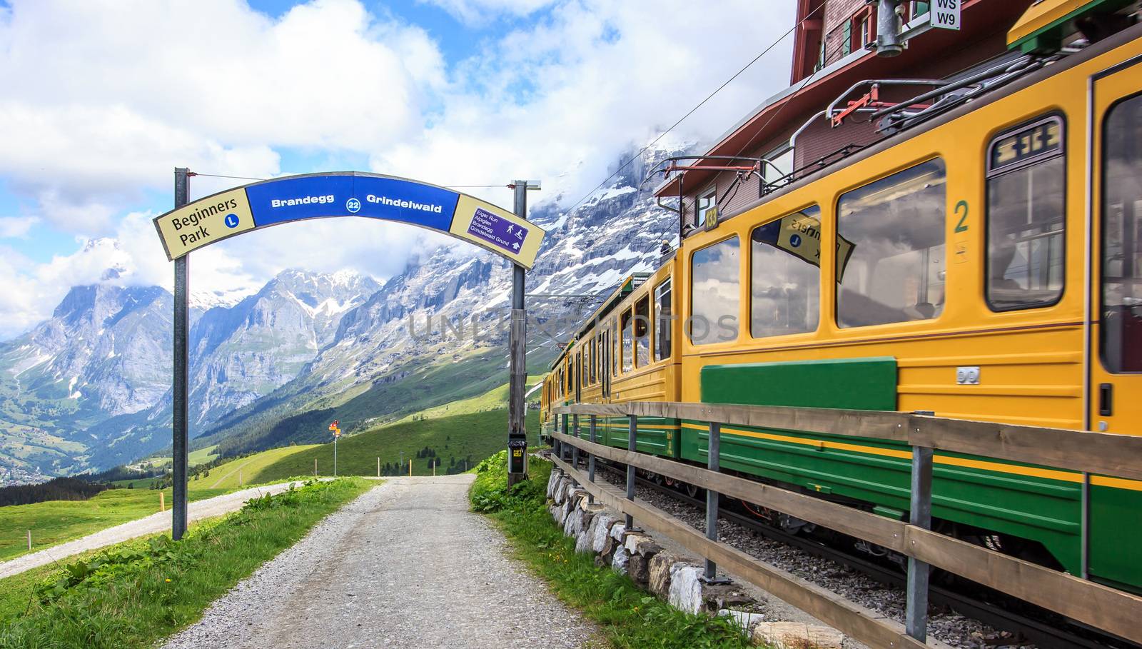 Sign of Starting point to begin walking trail with view of Swiss wengernalpbahn railways train departing Kleine Scheidegg station to Grindelwald, Switzerland. Walking trail along green fresh meadow with snow capped mountains as a background