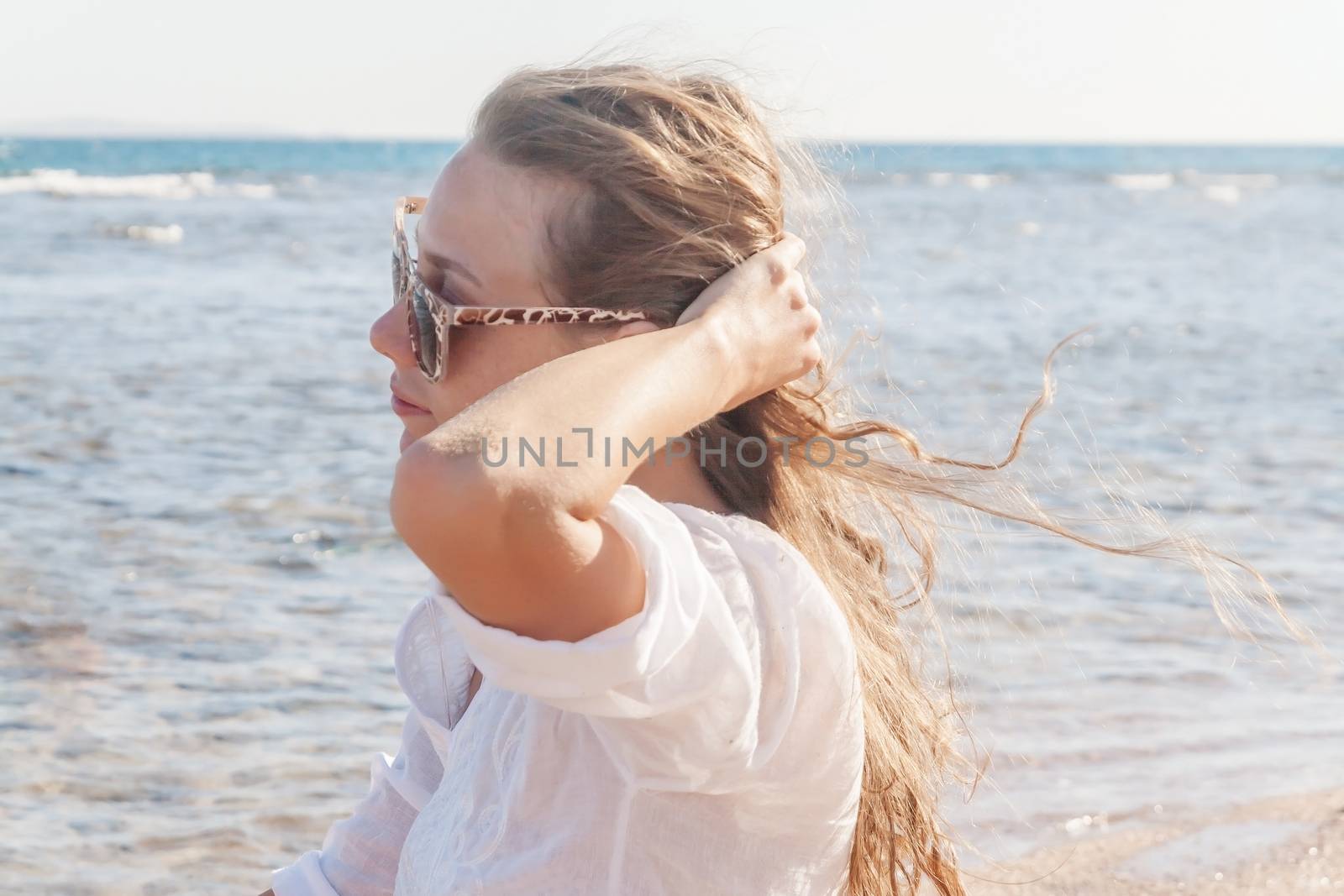 A young woman in sunglasses holds a hand to her hair fluttering in the wind near the sea. The concept of a free way of life.