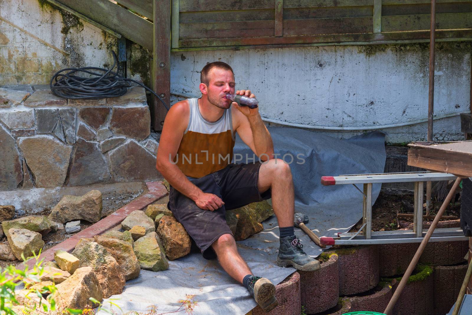 Craftsmen in work clothes takes a break. Sitting on the floor of a construction site holding a refreshment drink in hand.