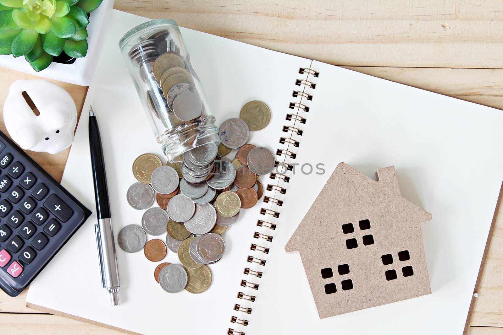 Business, finance, savings, property ladder or mortgage loan concept : Flat lay or top view of wood house model and coins scattered from glass jar on open blank notebook paper